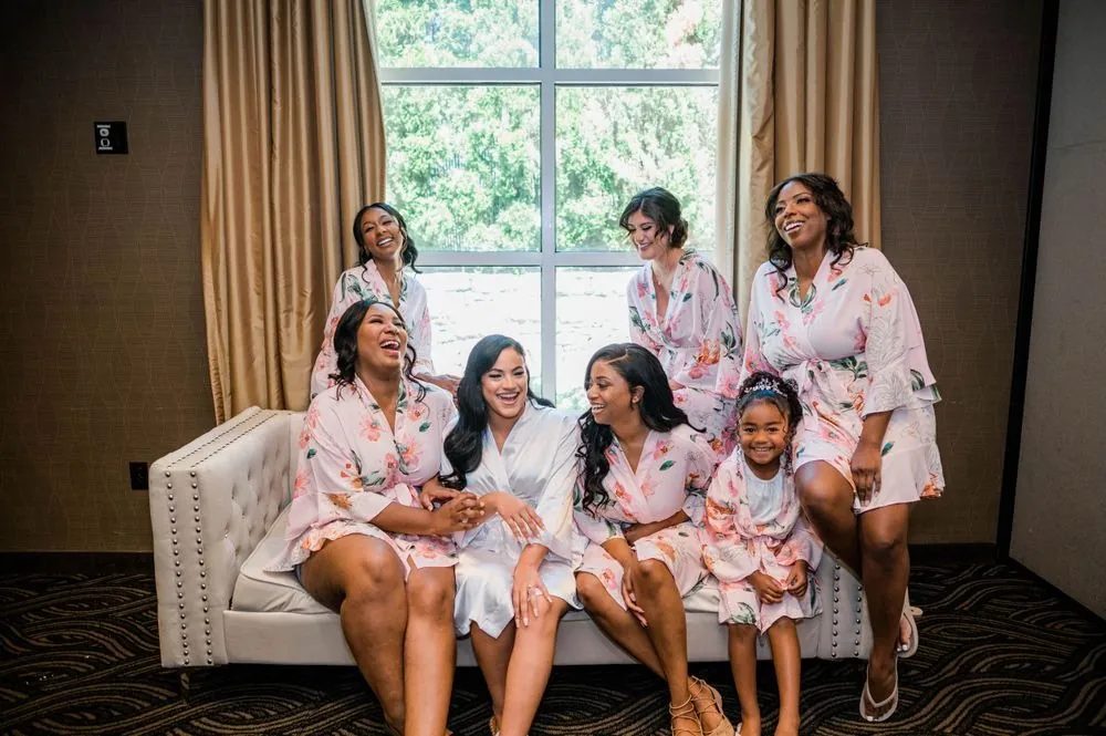 Bridesmaids in matching pink robes crowd onto a tufted sofa, laughing together in the bridal suite