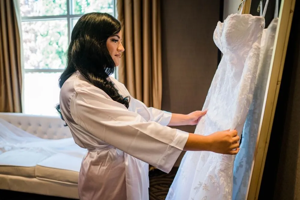 The bride in her robe reaches for the lace gown hanging by the window