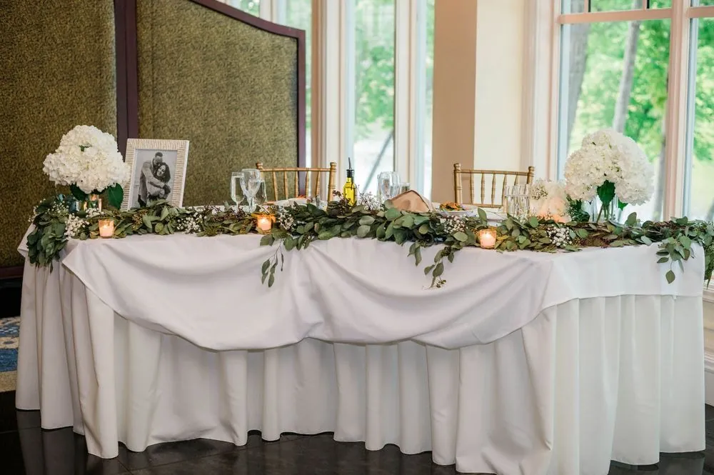 The head table dressed in white linens, candles and trailing greenery beside tall windows