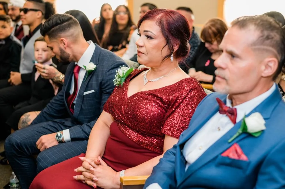 Guests in a red sequined dress and a blue suit watch the vows from their seats