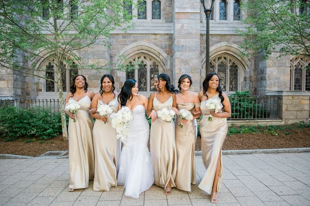 Bridesmaids in champagne satin gowns stand with the bride in front of Gothic stone windows