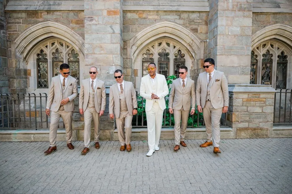 Groomsmen in tan suits and sunglasses line up with the groom outside a stone building