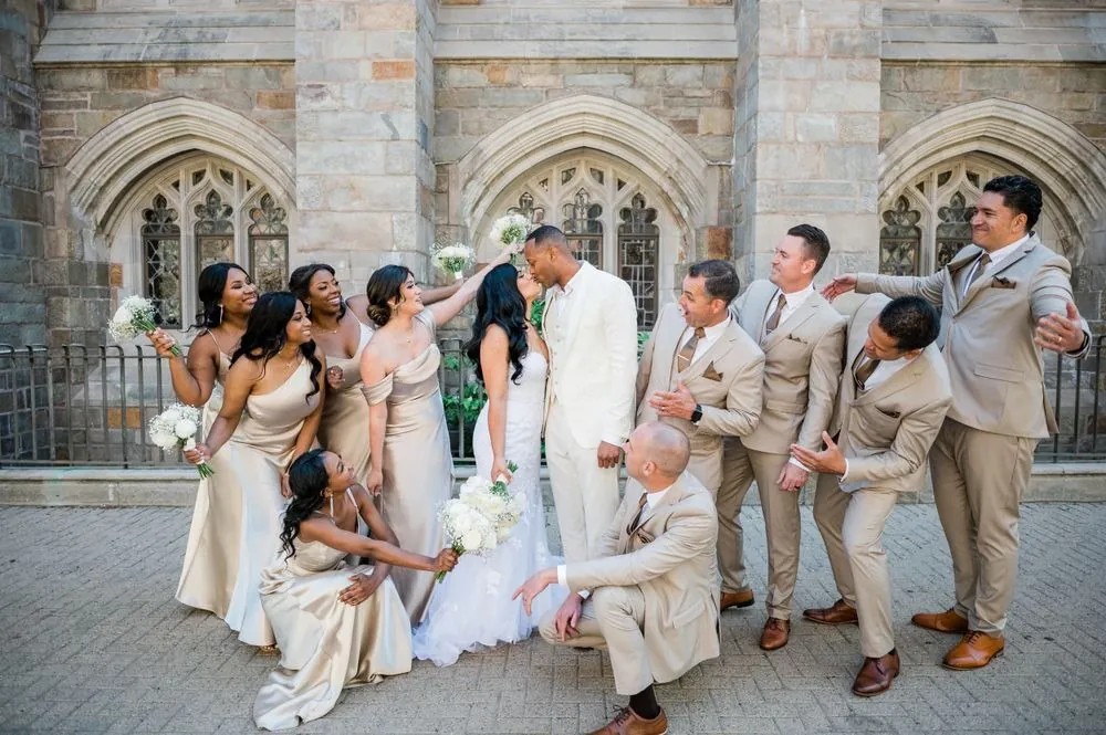 The full wedding party breaks into laughter and playful poses on a stone plaza