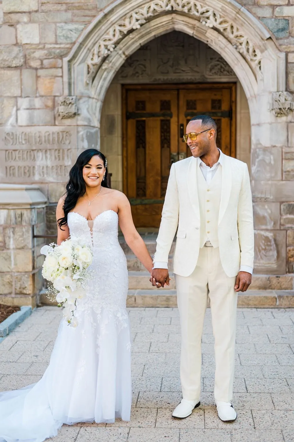 Sollisa and DJ stand hand in hand before an arched wooden door framed in carved stone