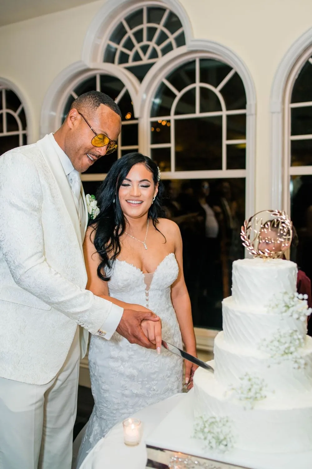 Sollisa and DJ cut their tiered white cake together beside arched windows