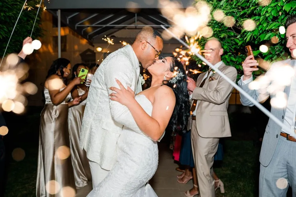 Sollisa and DJ kiss through a tunnel of sparklers as friends in satin and tan suits cheer