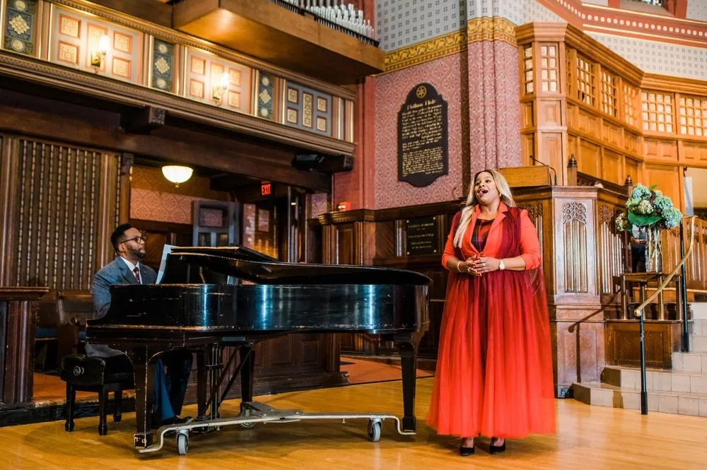 A singer in a red dress performs beside a grand piano below the organ pipes