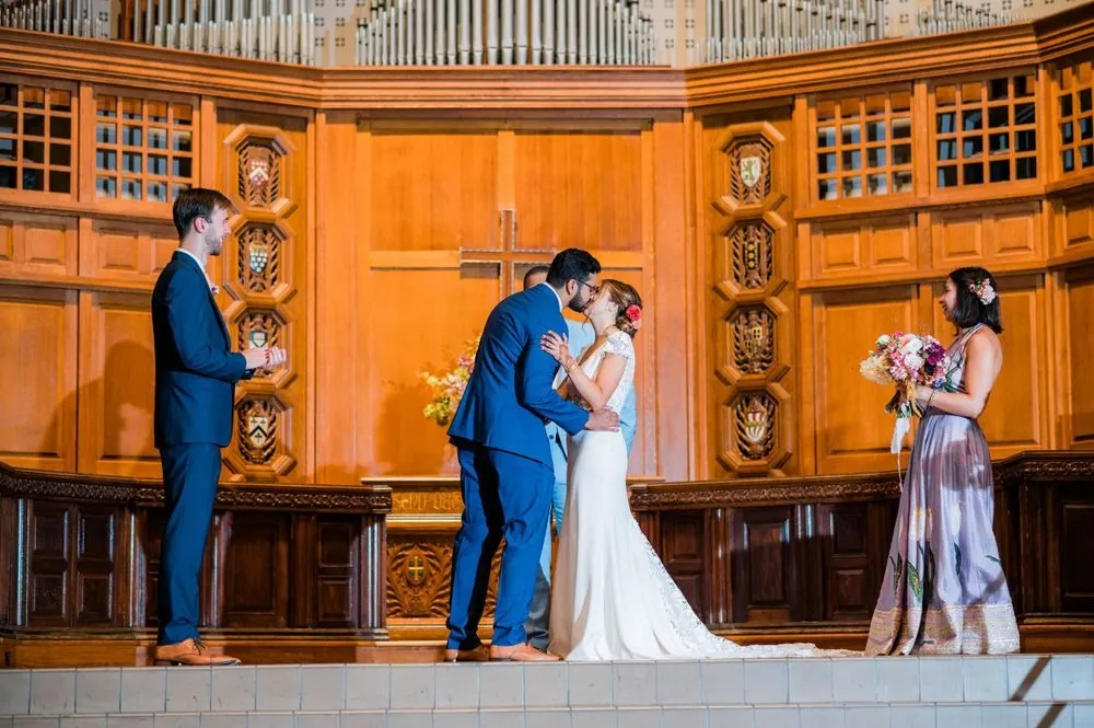 The couple kiss at the front of the chapel between their attendants and oak paneling