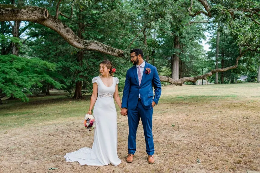 The couple hold hands on the grass under a sprawling tree branch in the park