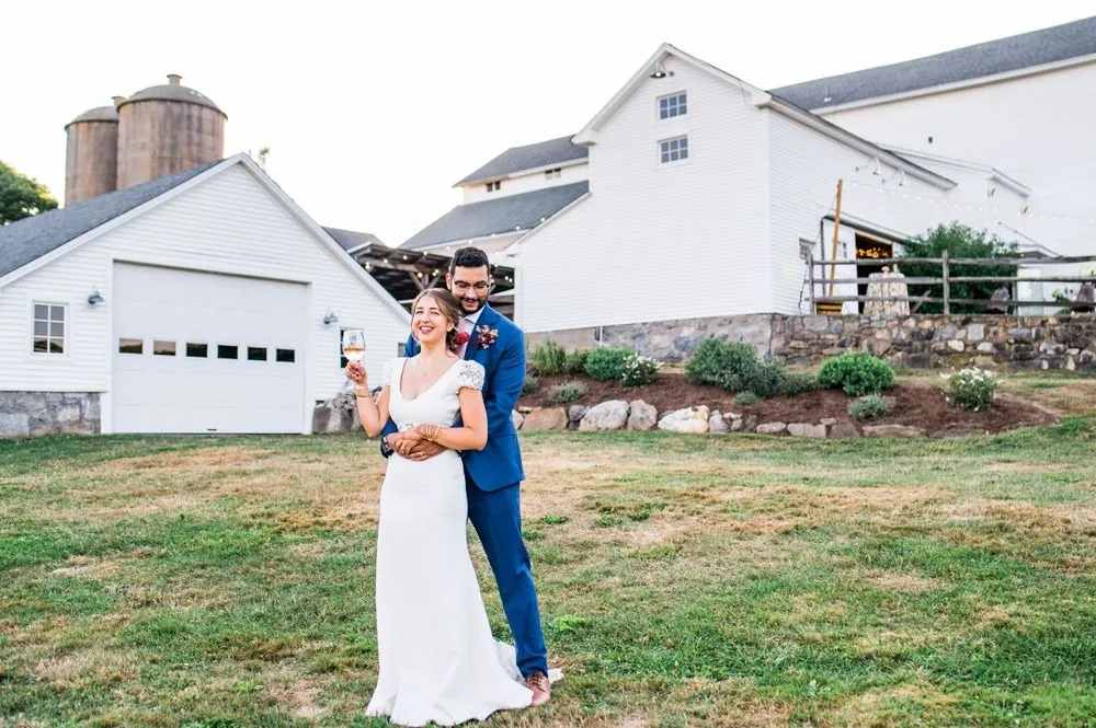 The groom hugs the bride from behind on the lawn with barns and silos behind them