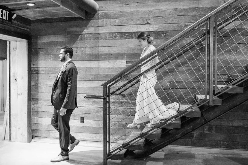 The bride walks down a steel staircase toward the groom in the barn, in black and white
