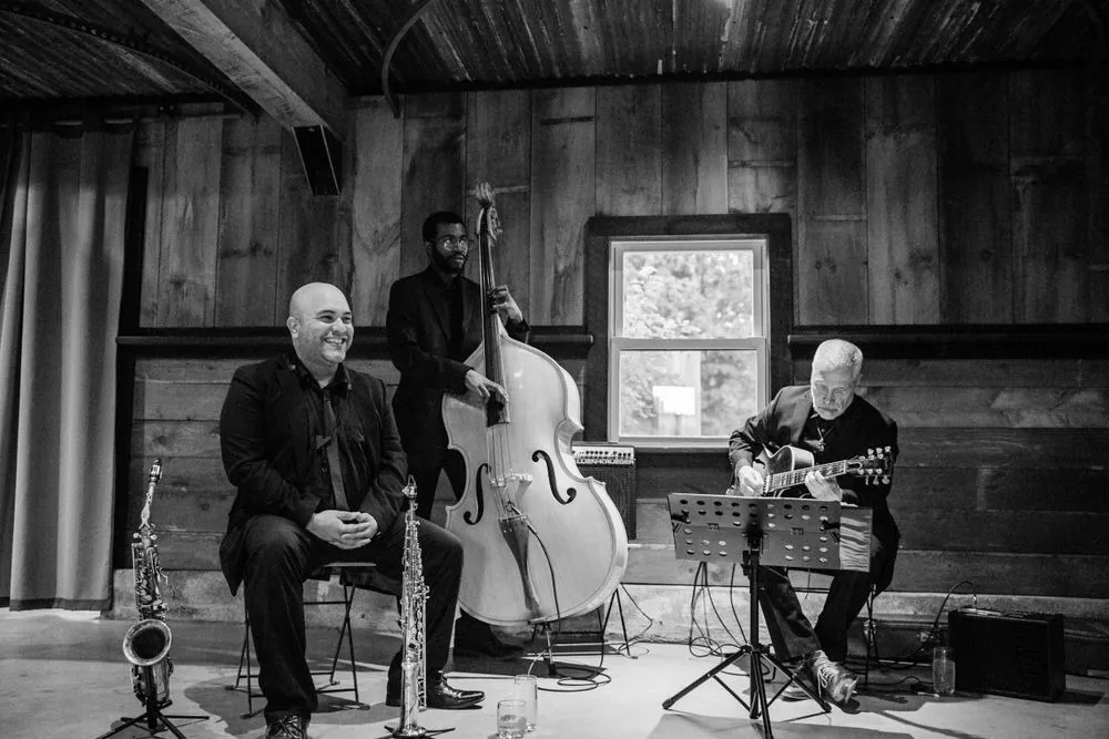 A trio on saxophone, upright bass, and guitar plays in the barn, in black and white