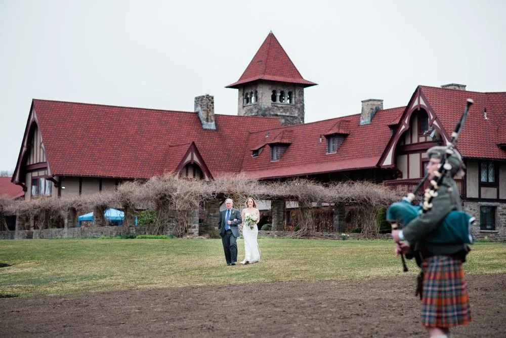 A bagpiper in a kilt plays on the lawn in front of the red-roofed castle