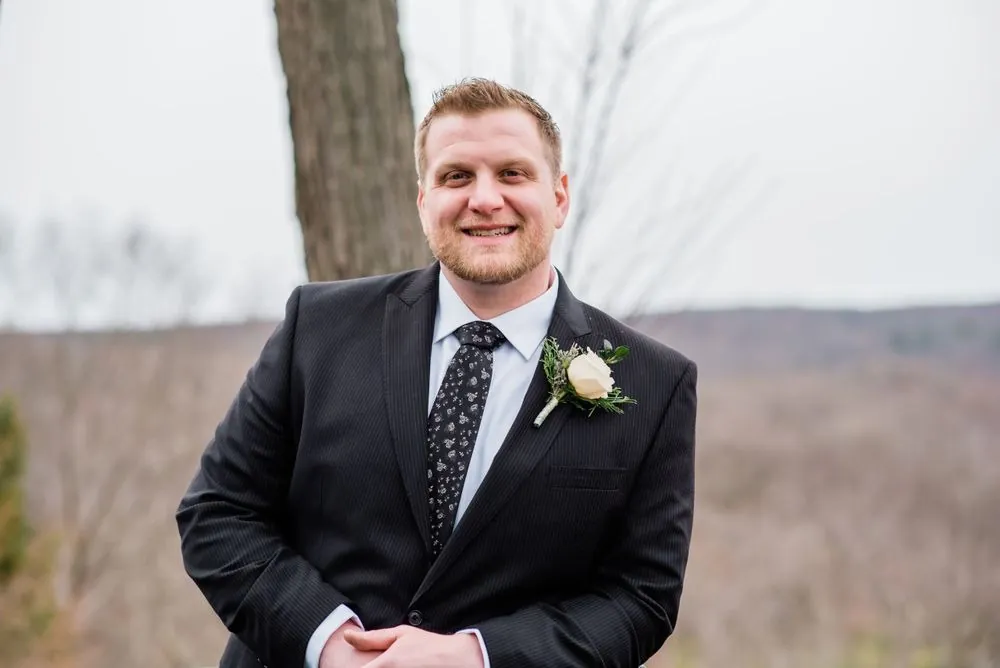 The groom grins in a dark suit with the bare hills behind him