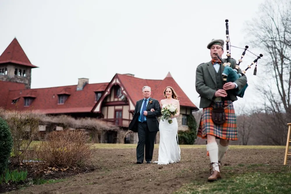 The bagpiper leads the bride and an escort across the lawn from the castle