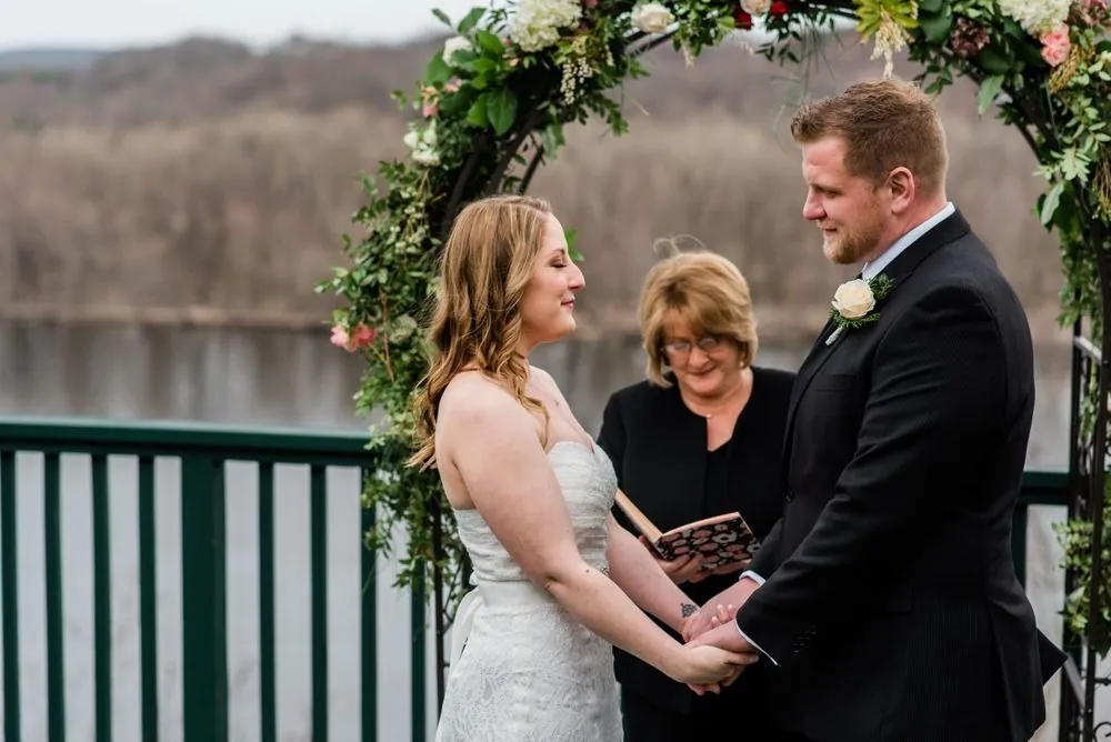 Taryn and Chris hold hands under the floral arch as the officiant reads