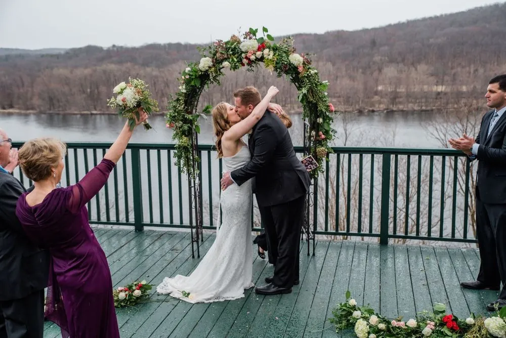 Taryn and Chris kiss under the rose arch as a guest in plum cheers, river behind them
