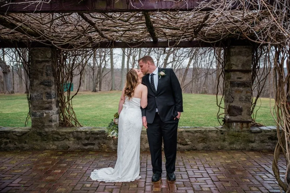 The couple kisses beneath the vine-covered stone pergola on a gray afternoon