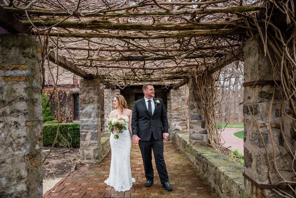 The couple stands together in the stone pergola under leafless twisting vines