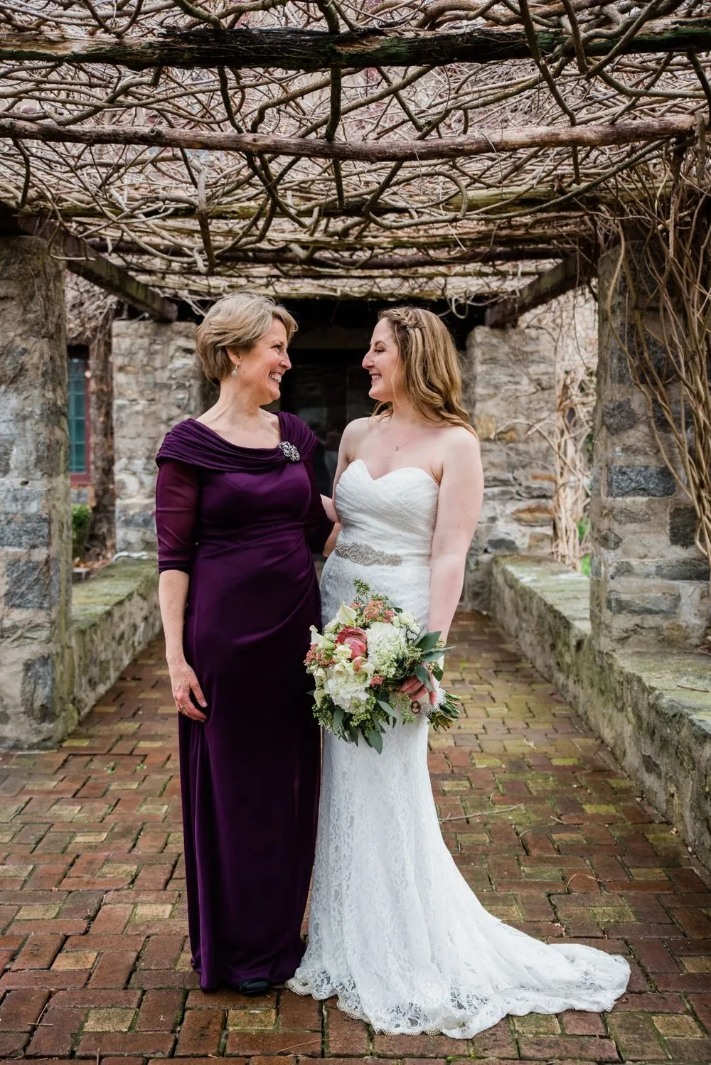 The bride stands with a woman in a plum dress in the pergola