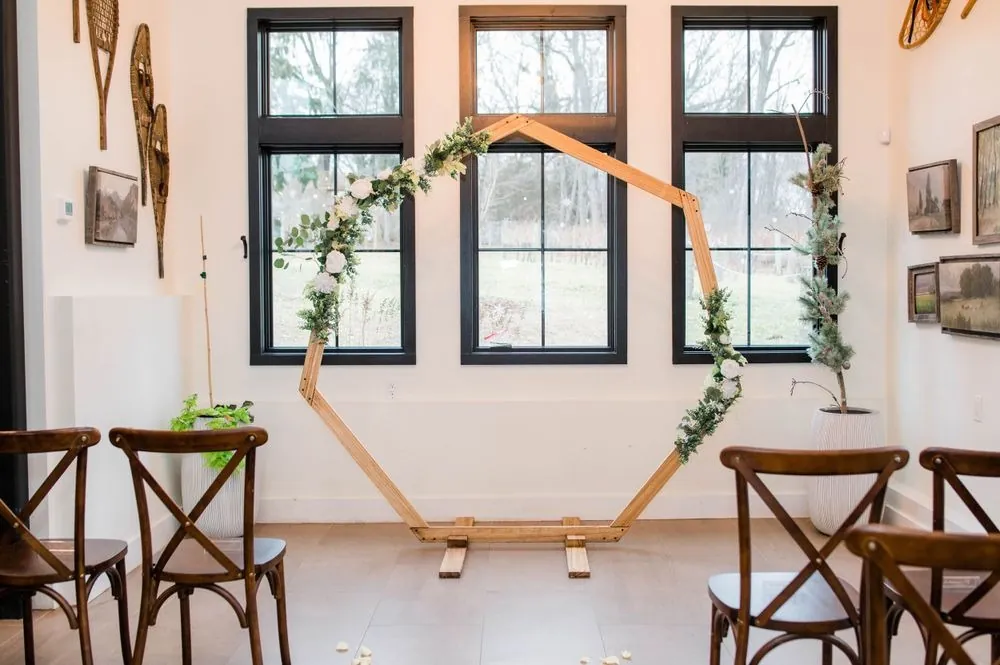 A wooden hexagon arch with white roses and greenery in front of black-framed windows
