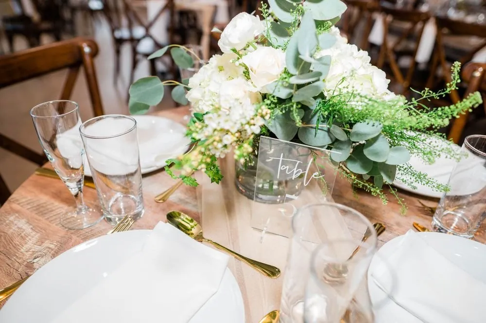 Round wood table set with white plates, gold flatware and a white rose centerpiece