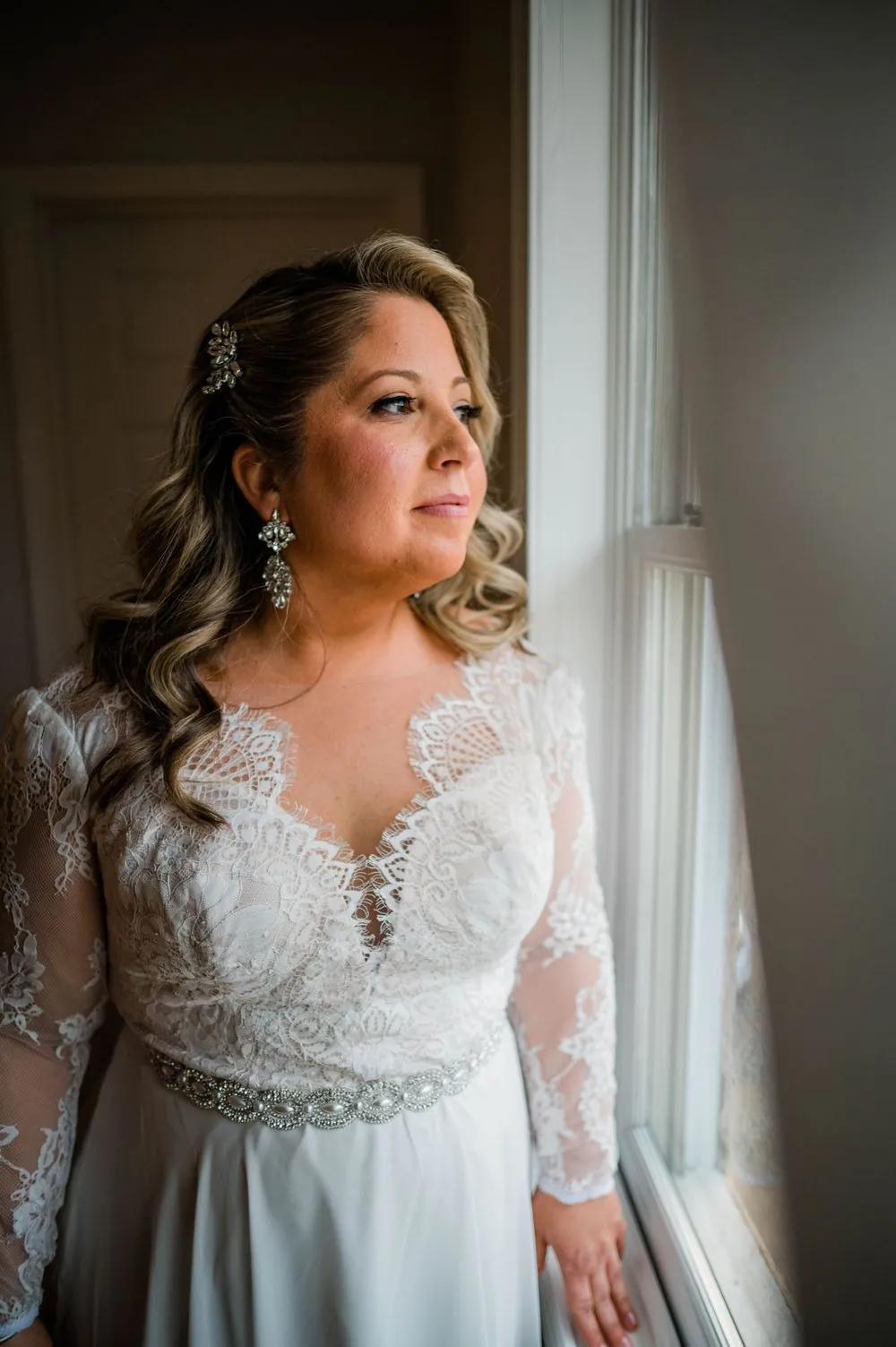 The bride in long lace sleeves looks out a tall window in soft daylight
