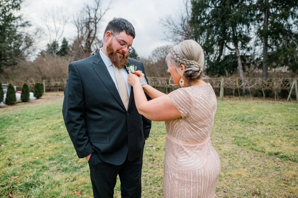 A woman in a sequined blush dress pins the groom's boutonniere outdoors