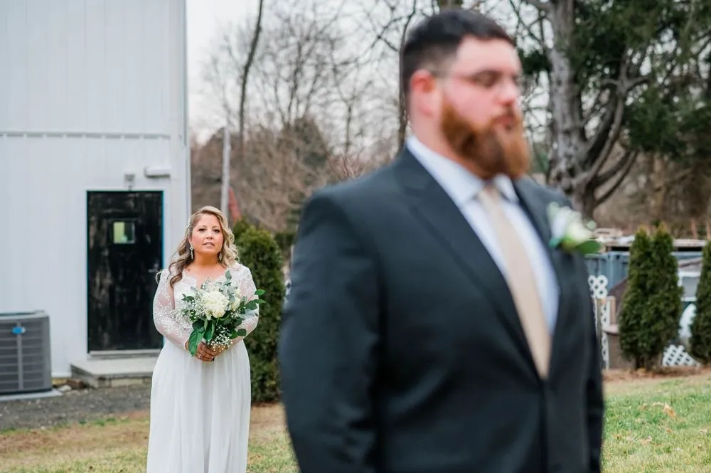 The bride walks up behind the groom for their first look beside a white building