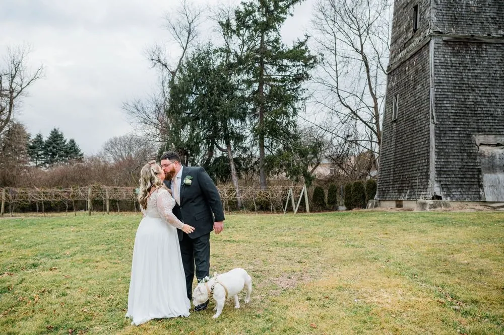 The couple kisses on the lawn beside a shingled tower as their bulldog sniffs the grass