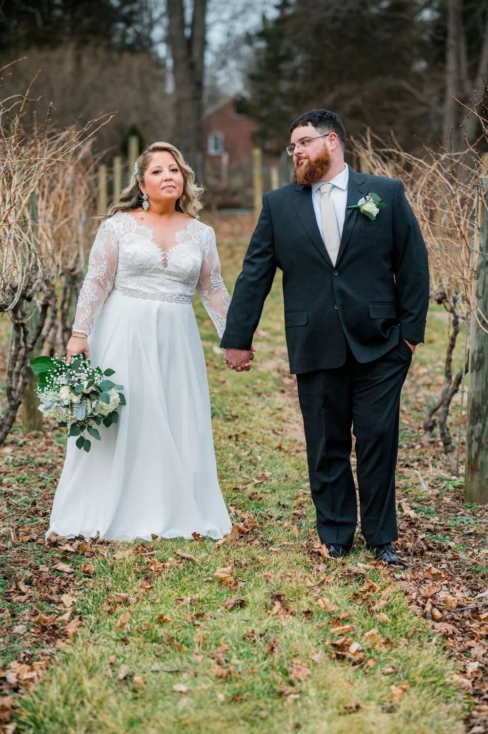 The couple stands among bare winter trees and dormant vines, holding hands