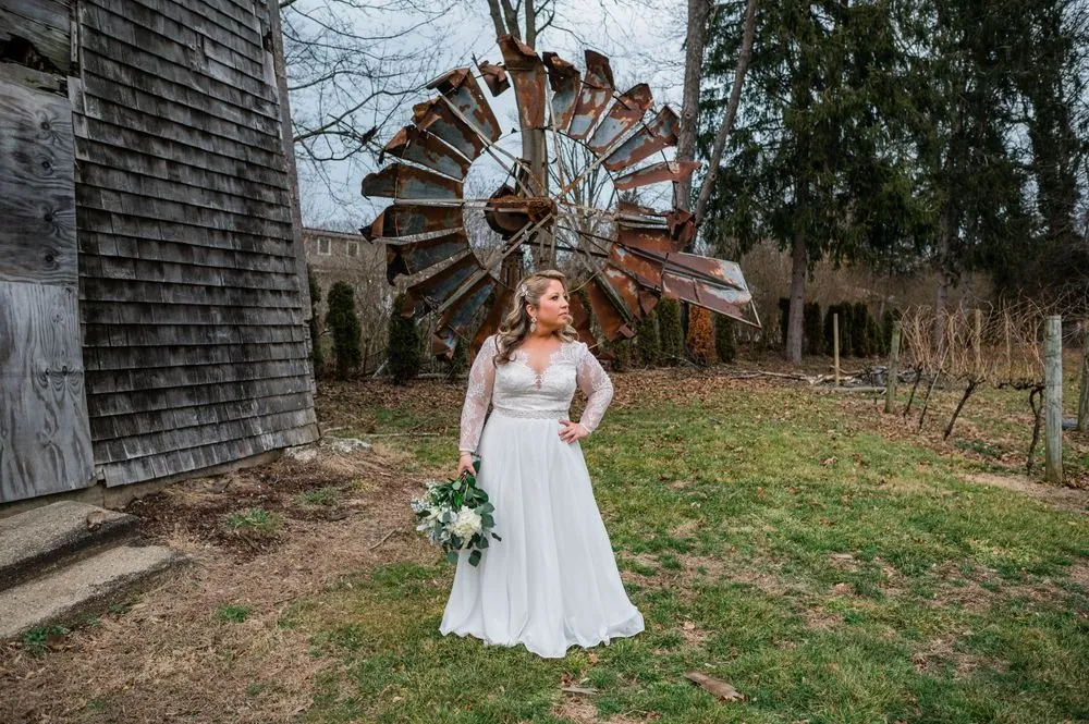 The bride stands before a rusted windmill and weathered shingles, bouquet at her side