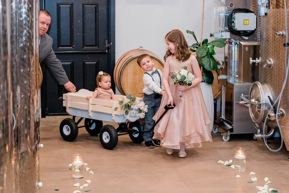 A flower girl pulls a toddler in a wooden wagon down the candlelit aisle between tanks