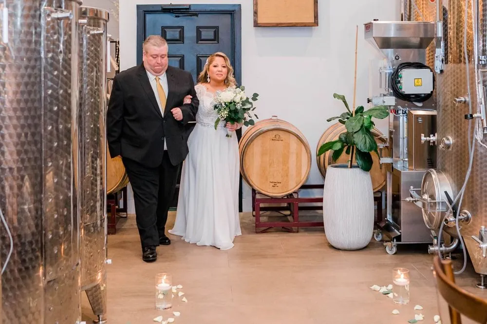 The bride walks in on a man's arm between steel tanks and a wine barrel