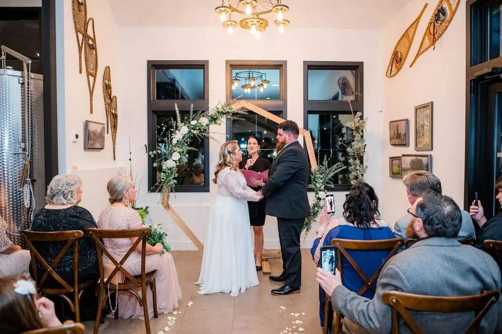 Guests on crossback chairs watch the vows beneath the arch in the bright tasting room