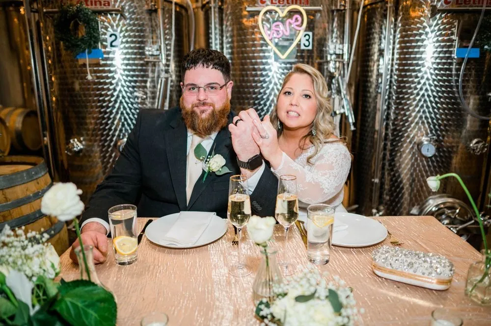 The couple holds up their ringed hands at the sweetheart table in front of steel tanks