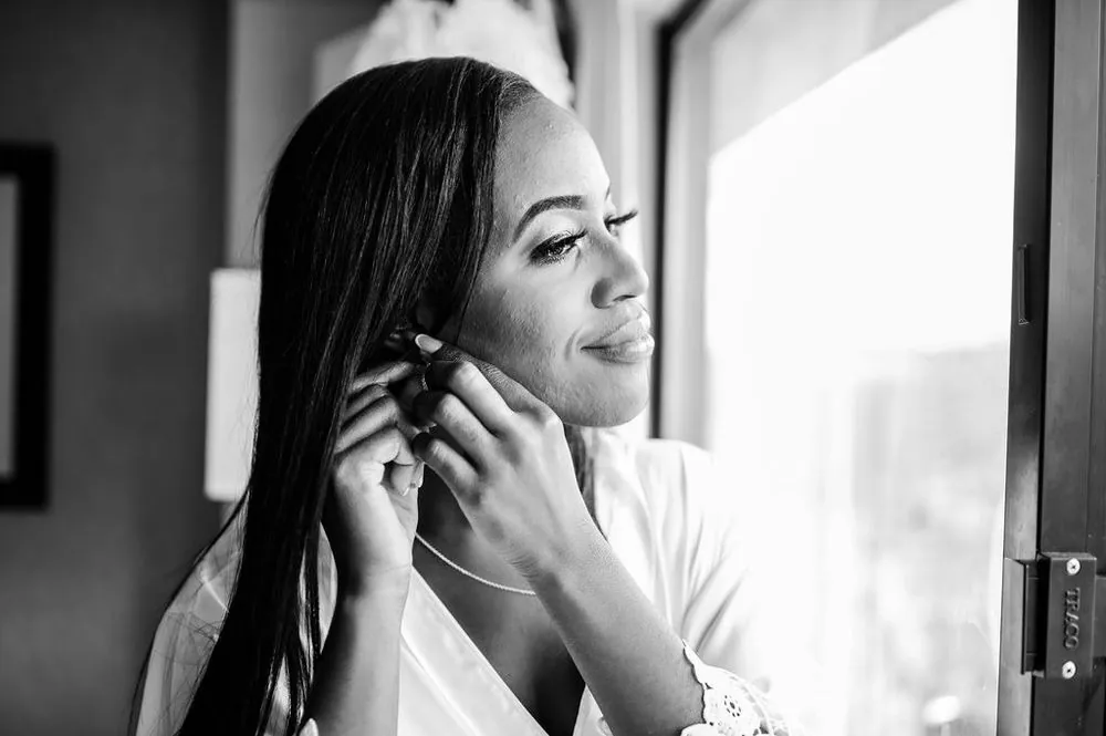 The bride fastens an earring by a bright window, smiling softly, in black and white