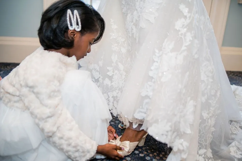 A little girl in a fuzzy white sweater kneels to fasten the bow on the bride's shoe