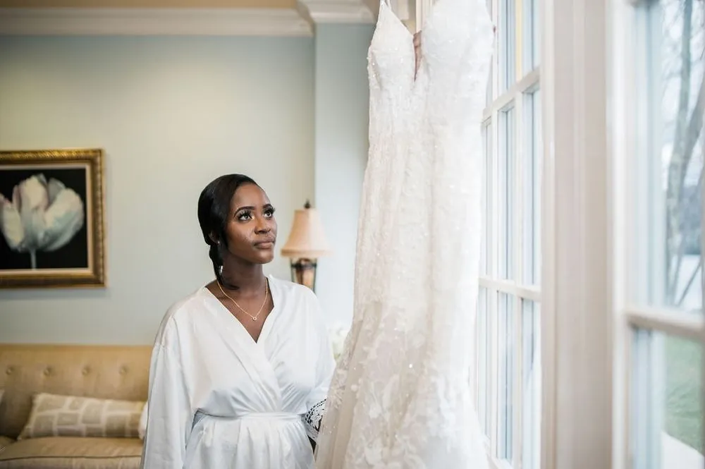 The bride in a white robe looks toward the window where her lace gown hangs