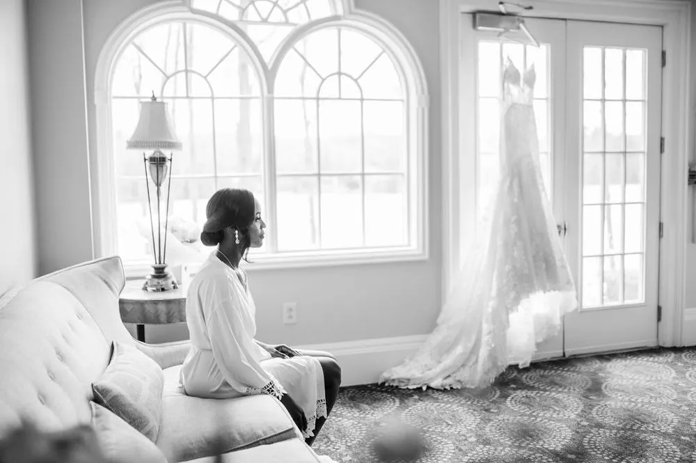 The bride sits quietly on a sofa below arched windows, her gown hanging nearby, in black and white