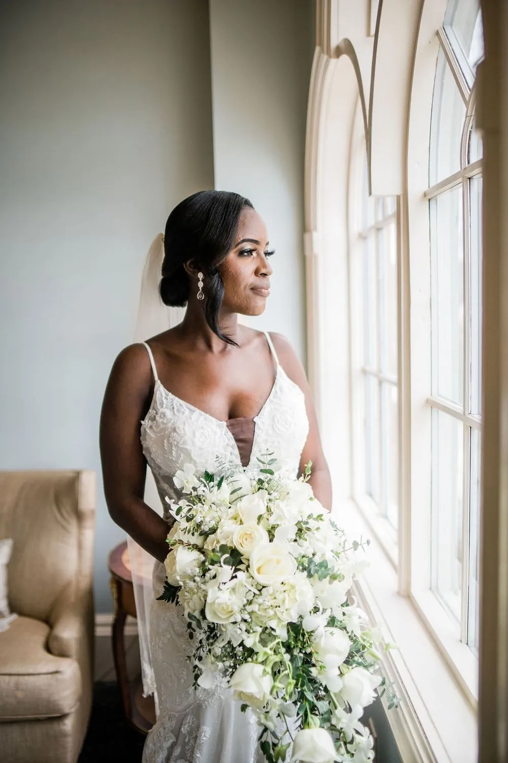 The bride holds a white bouquet and looks out a tall window in her lace gown