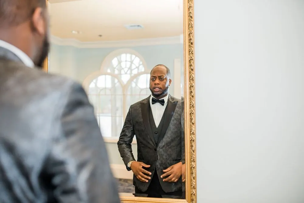 The groom checks his patterned jacket and bow tie in a gold-framed mirror