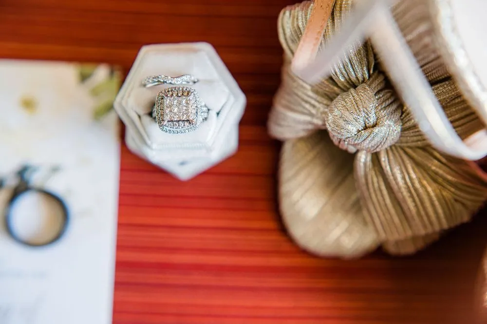 An engagement ring in a velvet hexagon box beside knotted gold shoes on red fabric