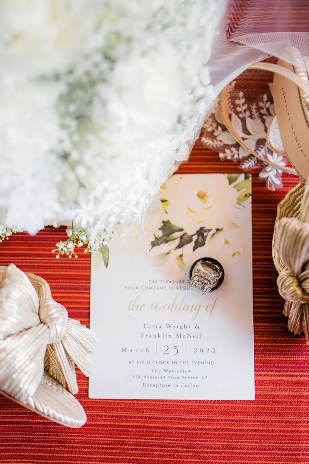 The wedding invitation and rings rest on red fabric beside white florals and gold shoes