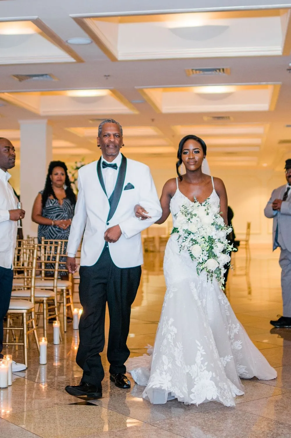 The bride walks the candlelit aisle on the arm of a man in a white dinner jacket