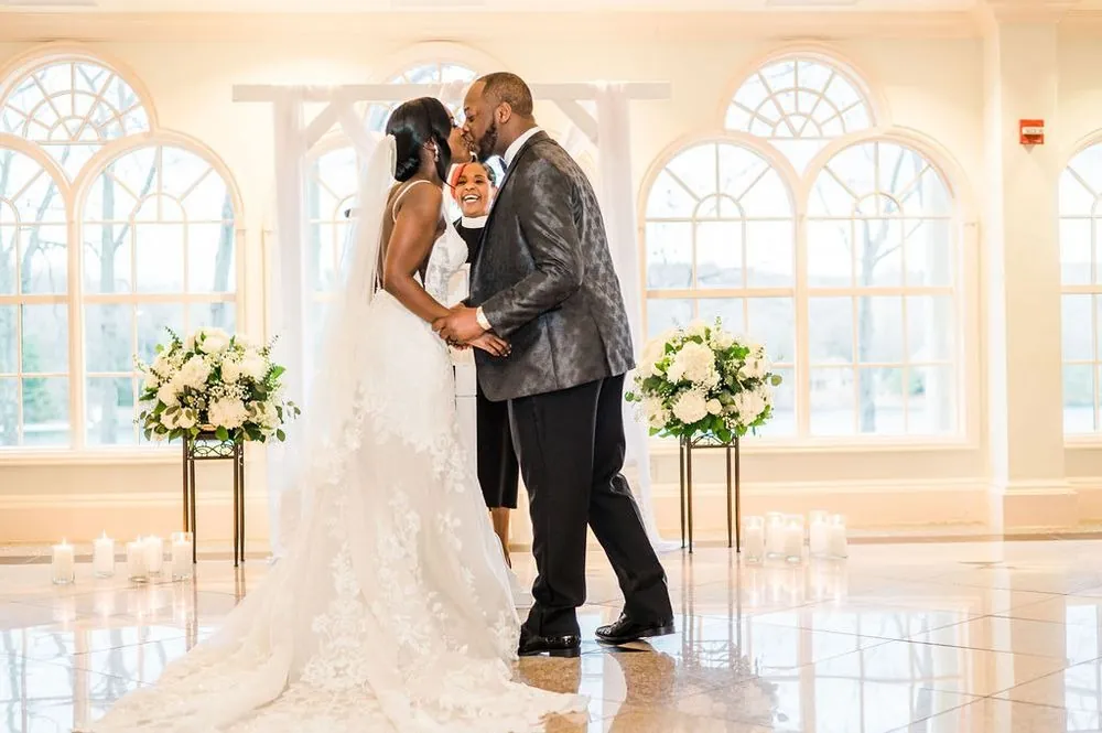 Taria and Franklin kiss between tall white floral arrangements in front of arched windows