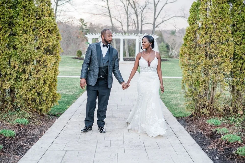 Taria and Franklin walk hand in hand down a garden path between tall evergreens