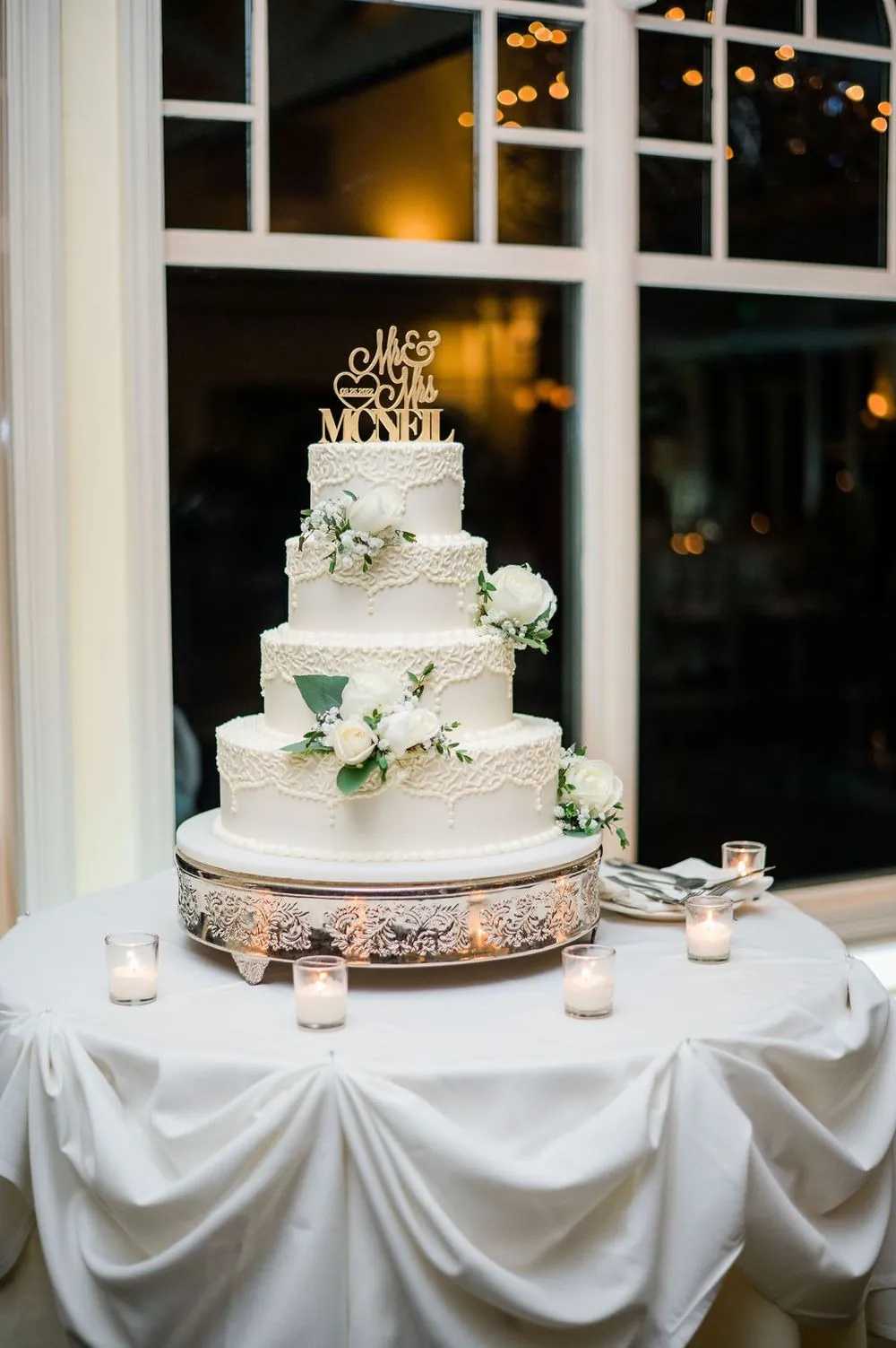 A tiered white cake with a gold topper stands before tall windows at dusk