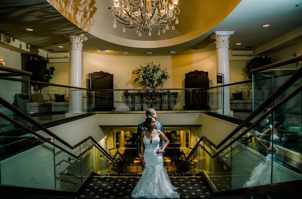 Taria stands on the grand staircase under a crystal chandelier with Franklin close behind her