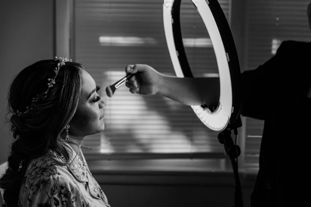 The bride has her makeup done beside a ring light, in black and white